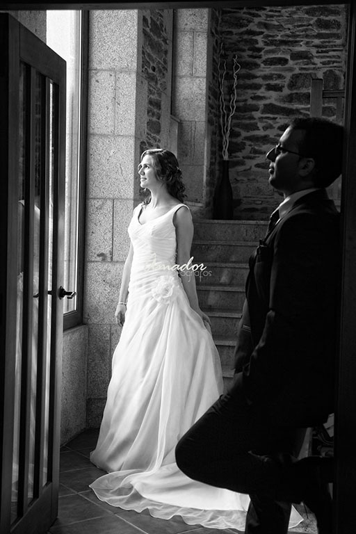 Foto en branco e negro dunha parella con traxes de boda mirando pola xanela.